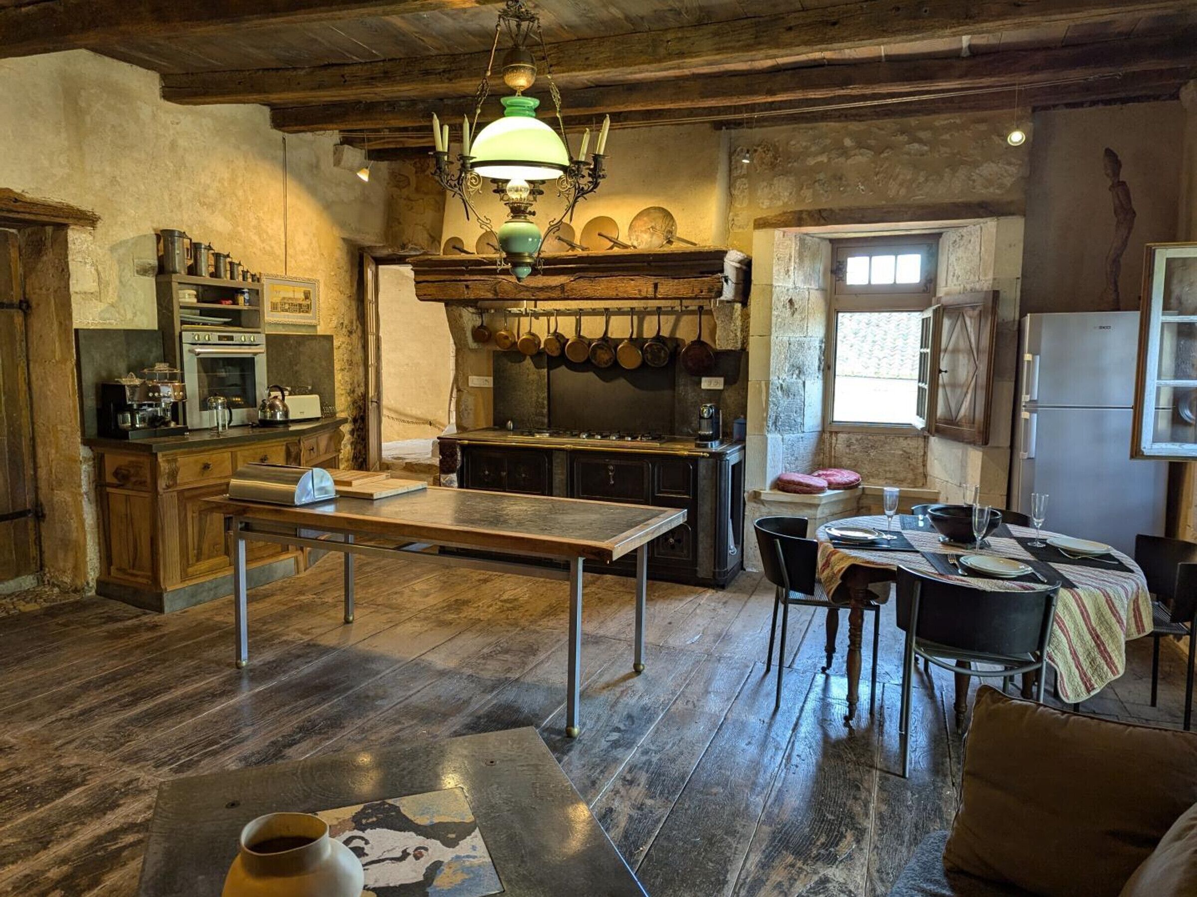 A rustic kitchen with wooden cabinets, a large island, hanging pots, and a vintage chandelier.