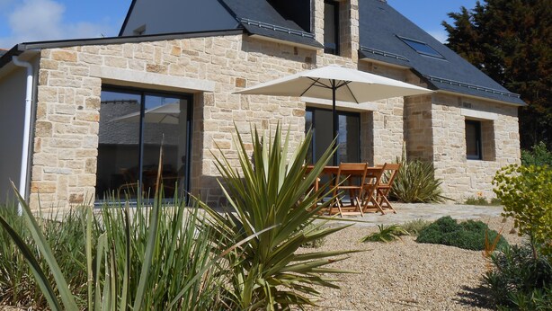 Une maison en pierre avec un toit en pente, de grandes fenêtres et une terrasse avec une table et des chaises.