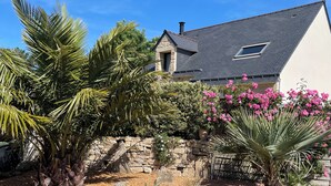 Une maison avec un mur en pierre, un escalier en pierre et un jardin agrémenté de palmiers et de fleurs roses.
