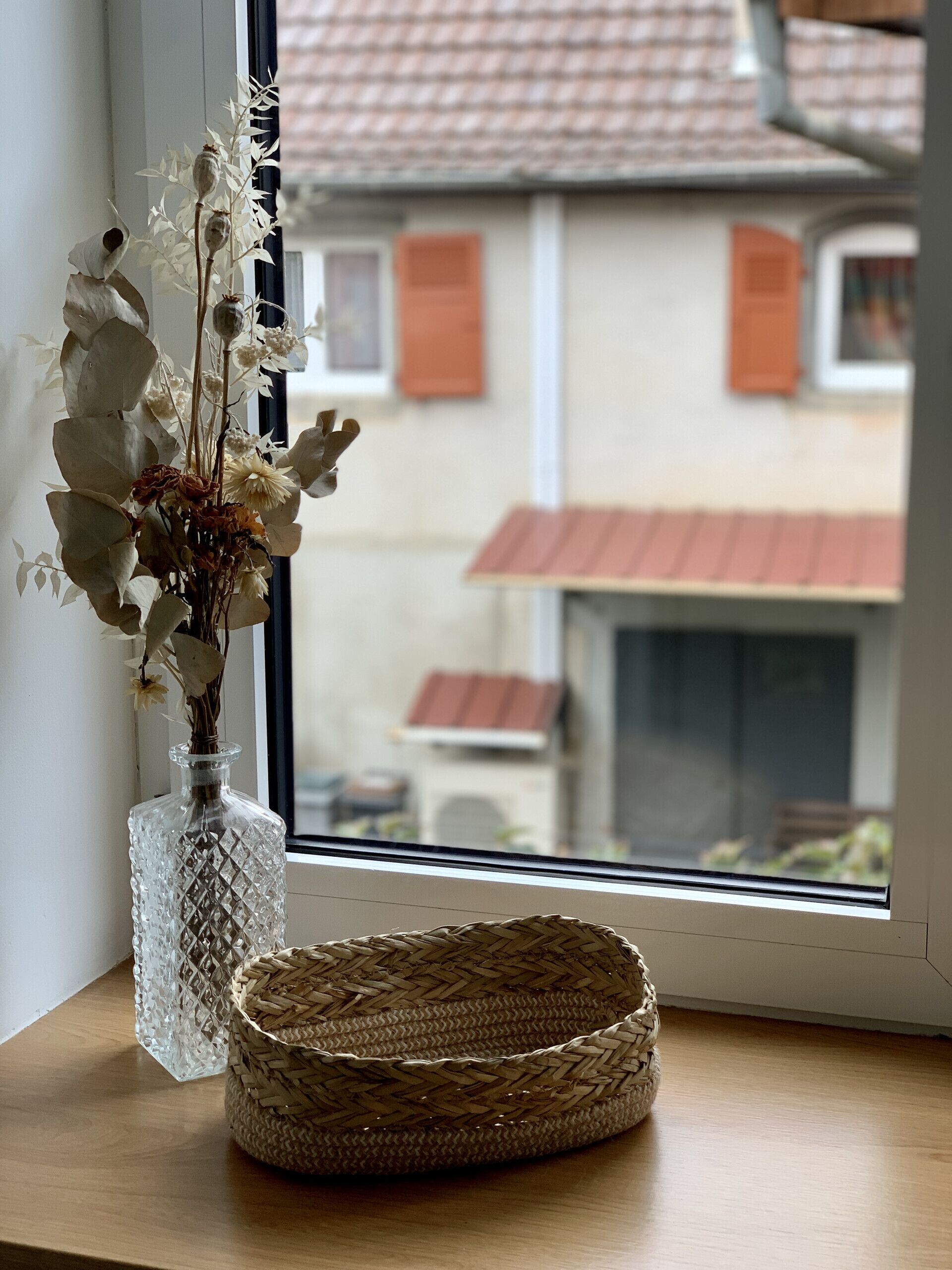 On voit clairement, à travers une fenêtre, un panier en osier et un vase en verre contenant des fleurs séchées, le tout posé sur une surface en bois.