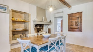 Un coin repas avec une table en bois, des chaises blanches dotées de coussins bleus, un luminaire suspendu et une étagère en pierre ornée d’objets décoratifs.