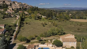 Un paysage rural avec une grande villa dotée d’une piscine, entourée de verdure et surplombant un village situé au loin.
