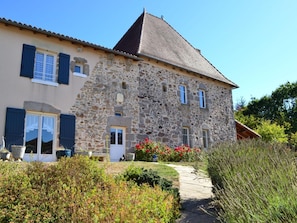 A stone house with a tiled roof, a front door, and a small garden with red flowers.