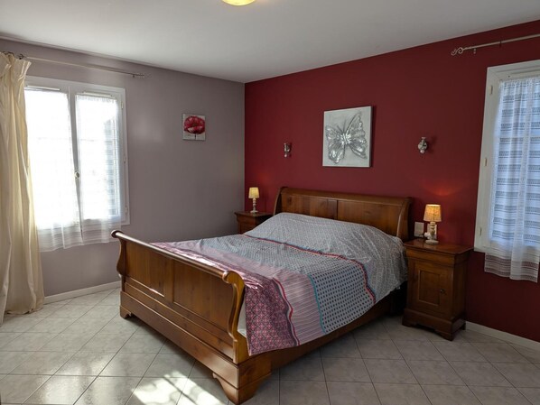 A bedroom with a wooden bed, two bedside tables with lamps, a window with curtains, and two wall-mounted light fixtures.