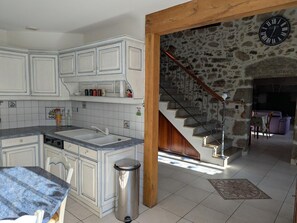 A kitchen with white cabinets, a sink, and a trash bin. There is a staircase leading to a living area with a stone wall and a clock.