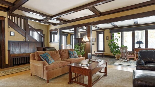 A spacious living room with a large sectional sofa, a wooden coffee table, and a staircase with dark wooden beams. - Historic Cozy Home Walkable to DT w/ Pool Table (Kalispell)