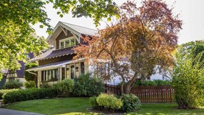 A two-story house with a covered porch, a well-maintained lawn, and a variety of trees and shrubs. - Historic Cozy Home Walkable to DT w/ Pool Table (Kalispell)