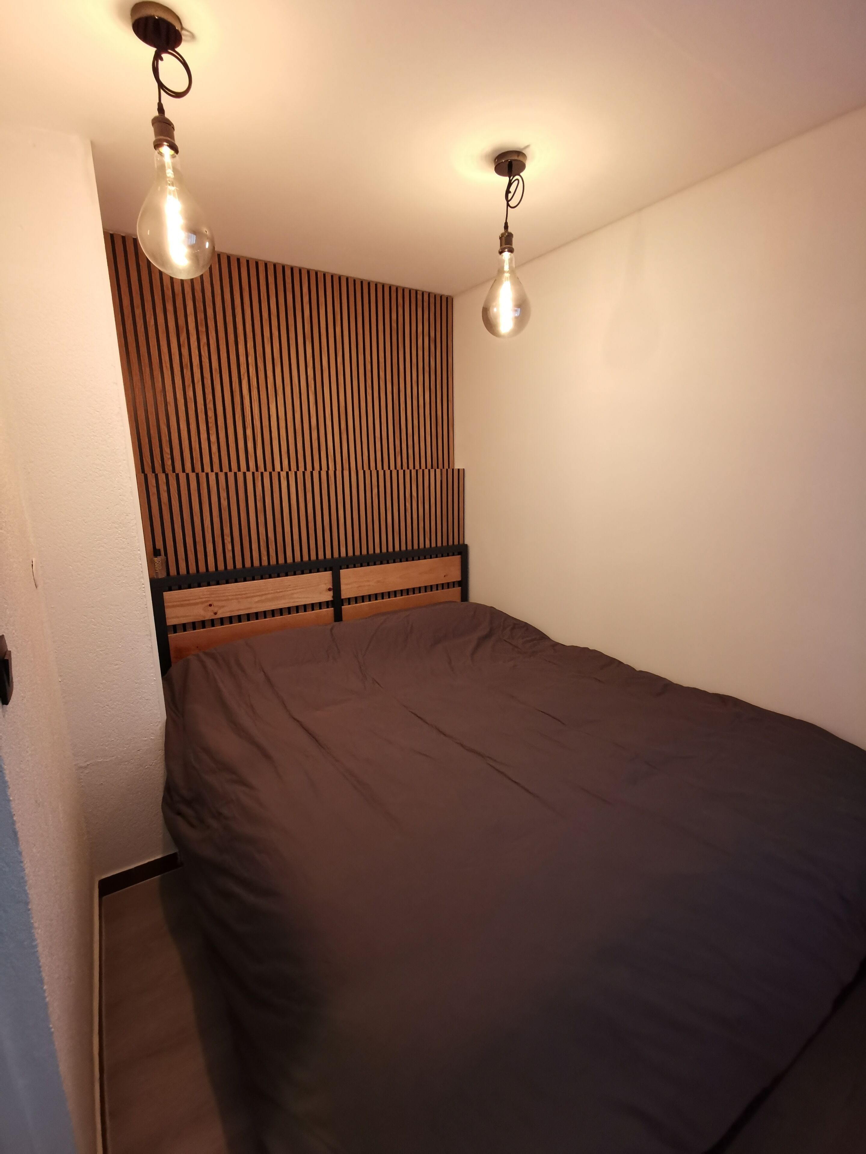 Quarto com cabeceira de ripas de madeira, uma cama de solteiro com colchão escuro e duas luminárias suspensas.