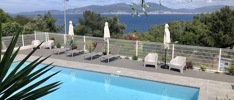 A pool area with lounge chairs and umbrellas, surrounded by a white fence, overlooking a coastal city and mountains.