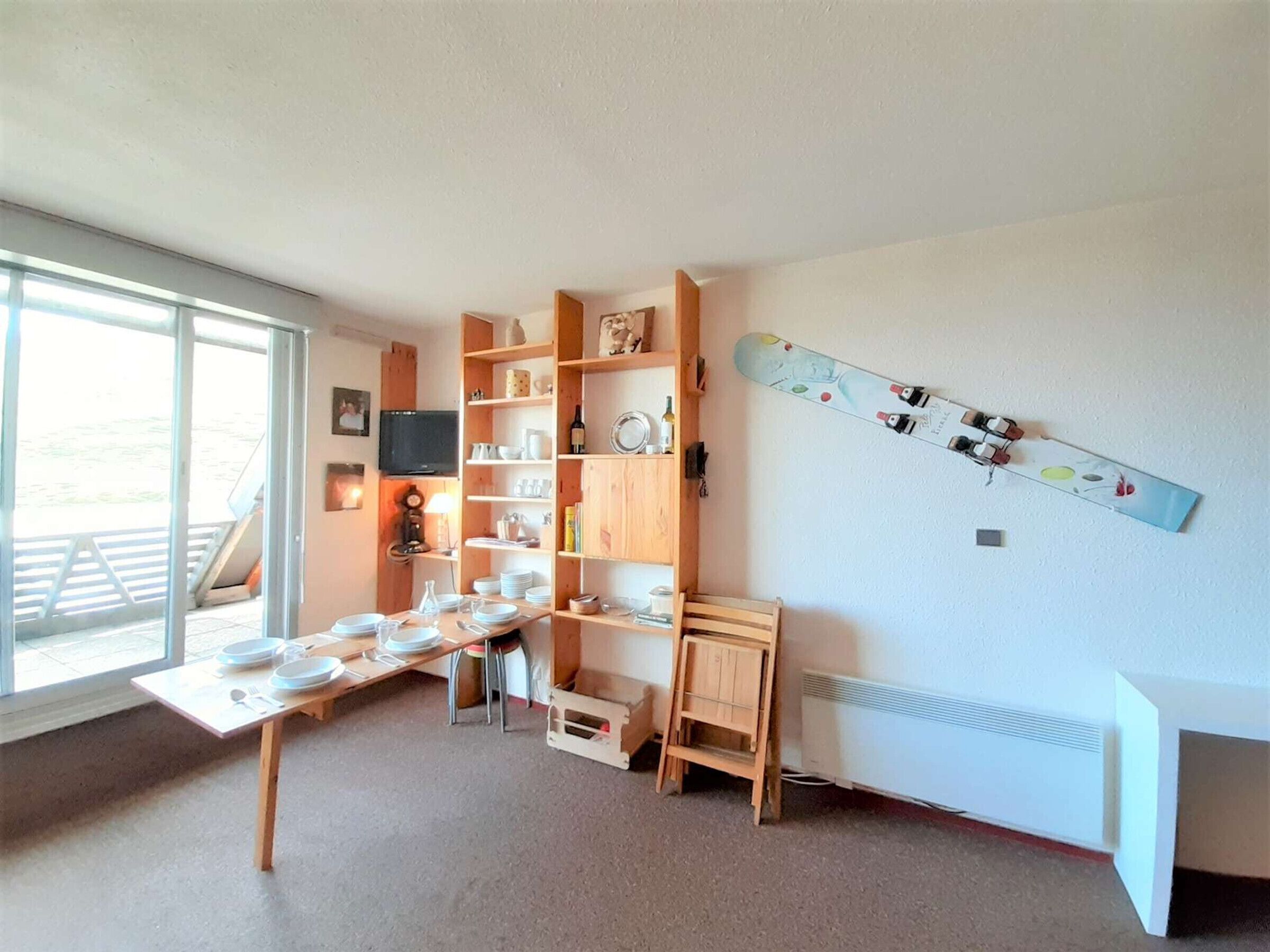 A room with a dining table set for four, a wooden bookshelf, a mounted snowboard, and a large window.