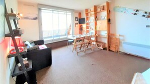 A spacious living room with a large window, a dining area with wooden chairs, and a built-in bookshelf.