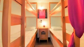 A small, narrow room with two bunk beds and a single bed. There is a small table with a lamp and a red curtain.
