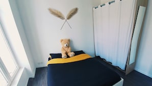 Een bed met een donkerblauwe en gele sprei, een teddybeer op het bed, een geweven vloerkleed en een raam met gordijnen.