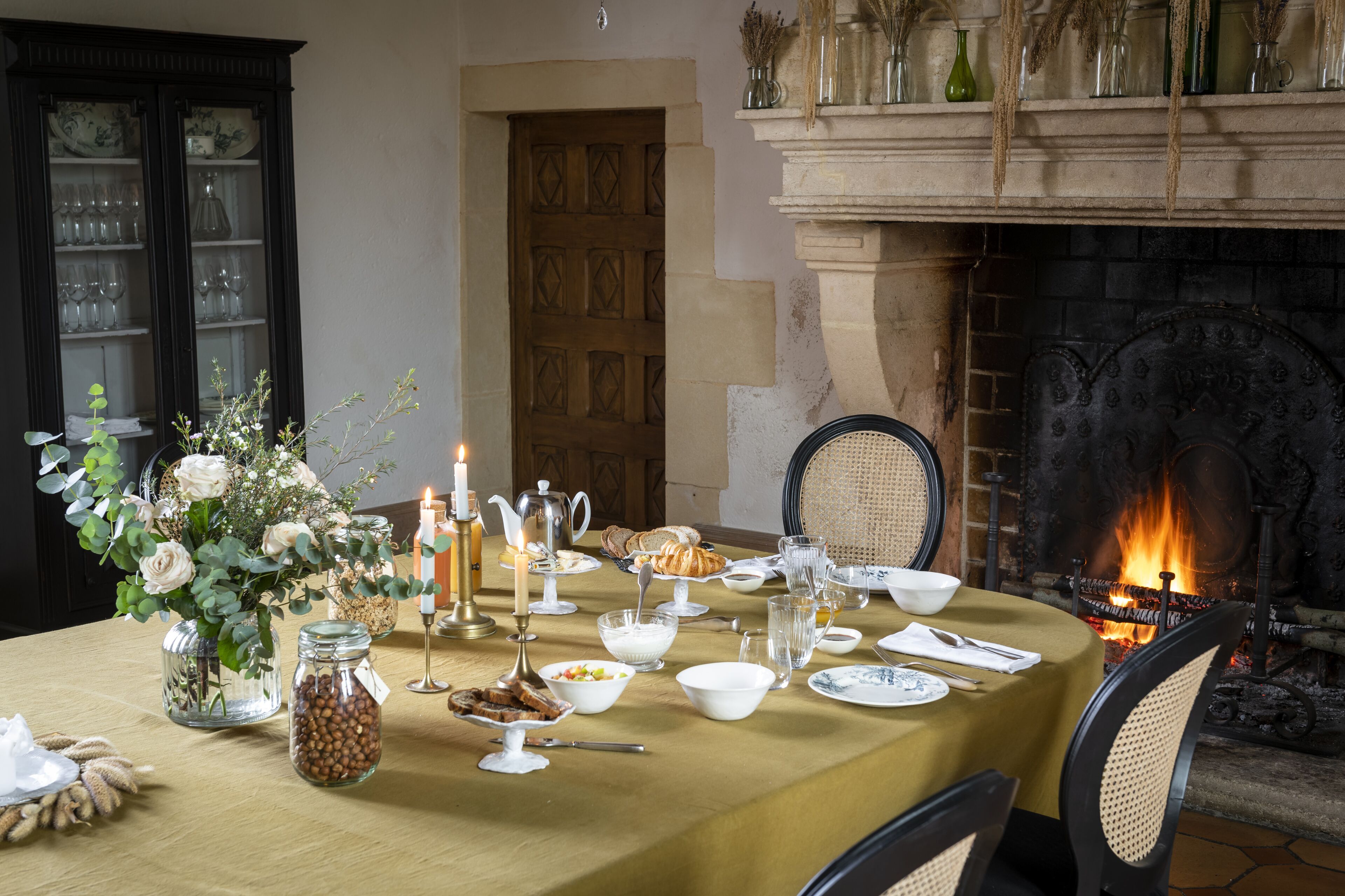 A dining table set for a meal with candles, a fireplace, and a view of the fire.