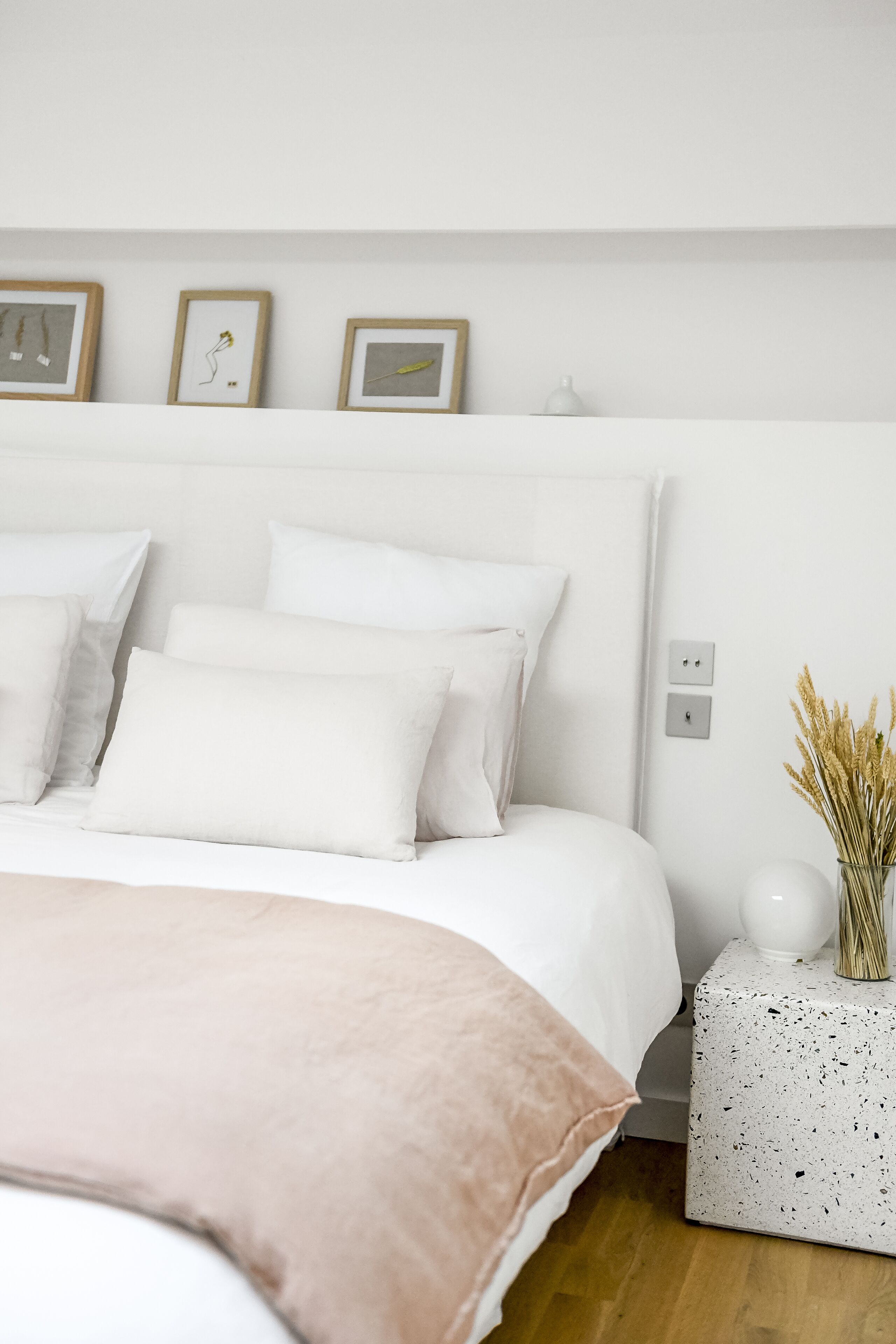 Ein ordentlich eingerichtetes Schlafzimmer mit einem großen Bett, weißer Bettwäsche und einem kleinen Nachttisch mit einer Vase getrockneter Blumen.