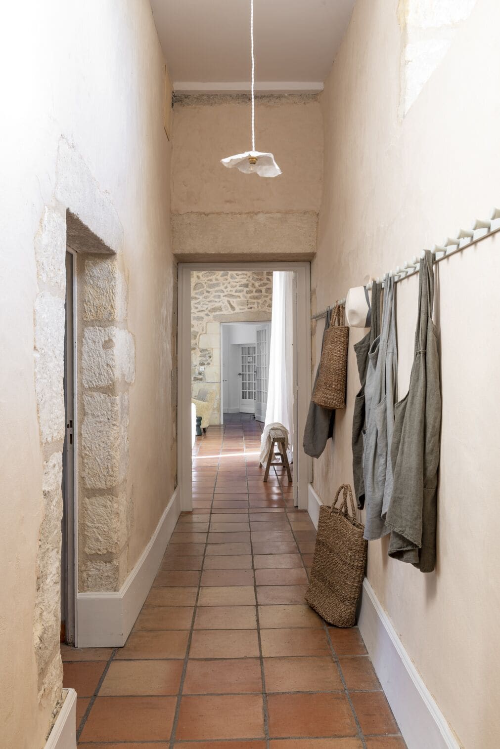 Un estrecho pasillo con una pared de piedra a un lado y suelo de baldosas. Ropa colgada en la pared y una cesta de mimbre en el suelo.