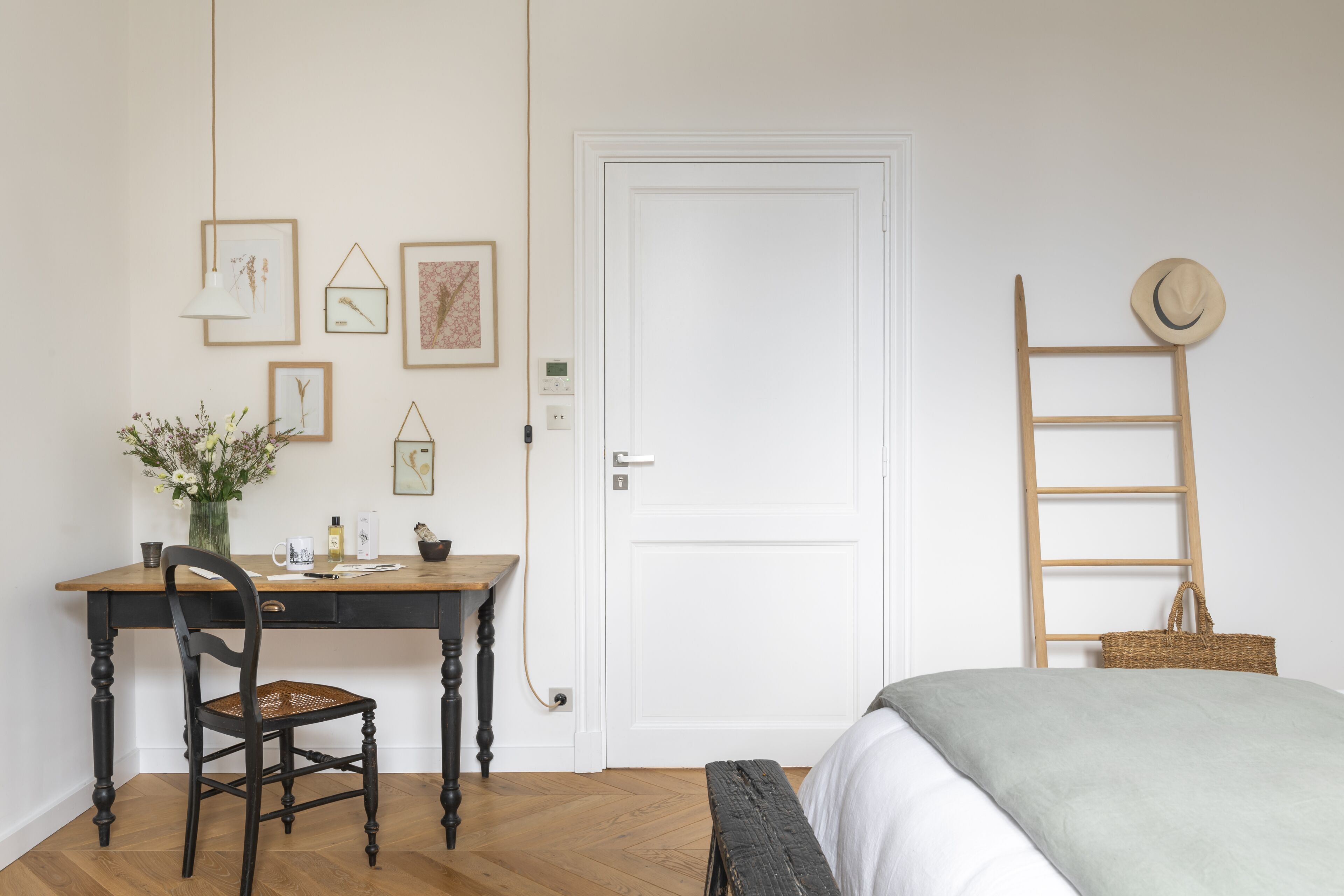 Una habitación con un escritorio de madera, una silla, una cama con una manta verde, una puerta blanca y una pared con cuadros enmarcados.