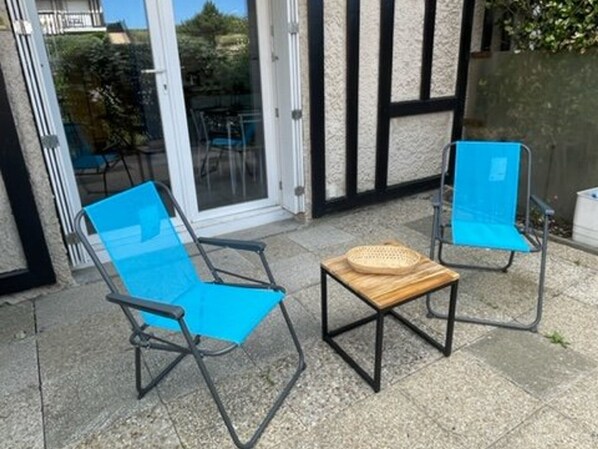 Dos sillas plegables azules y una pequeña mesa de madera con una cesta tejida en un patio con una puerta de cristal que da a un jardín.