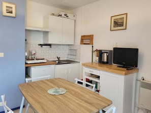 A compact kitchen with white cabinets, a wooden table, a coffee maker, a kettle, and a television.