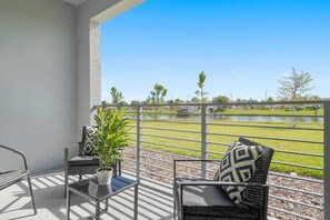 A balcony with a view of a grassy area and a body of water. There is a table with a potted plant and two chairs with patterned cushions.