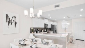 Una cocina moderna con armarios blancos, una isla central y un comedor preparado para seis personas, con sillas blancas y una araña.