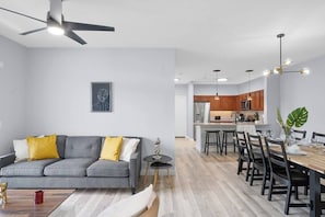 Una sala de estar moderna con un sofá gris, cojines amarillos, una mesita redonda y un ventilador de techo.