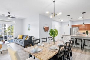 Una cocina moderna con una isla central, un comedor con mesa de madera dispuesta para seis personas, y una sala de estar con sofá y un ventanal grande.