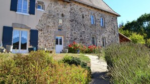 A stone house with a tiled roof, a front door, and a small garden with red flowers.