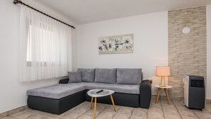 A modern living room with a grey sectional sofa, a small round table with a remote control, a standing lamp, and a wall-mounted air conditioner. - Apartment Susa (Vir)