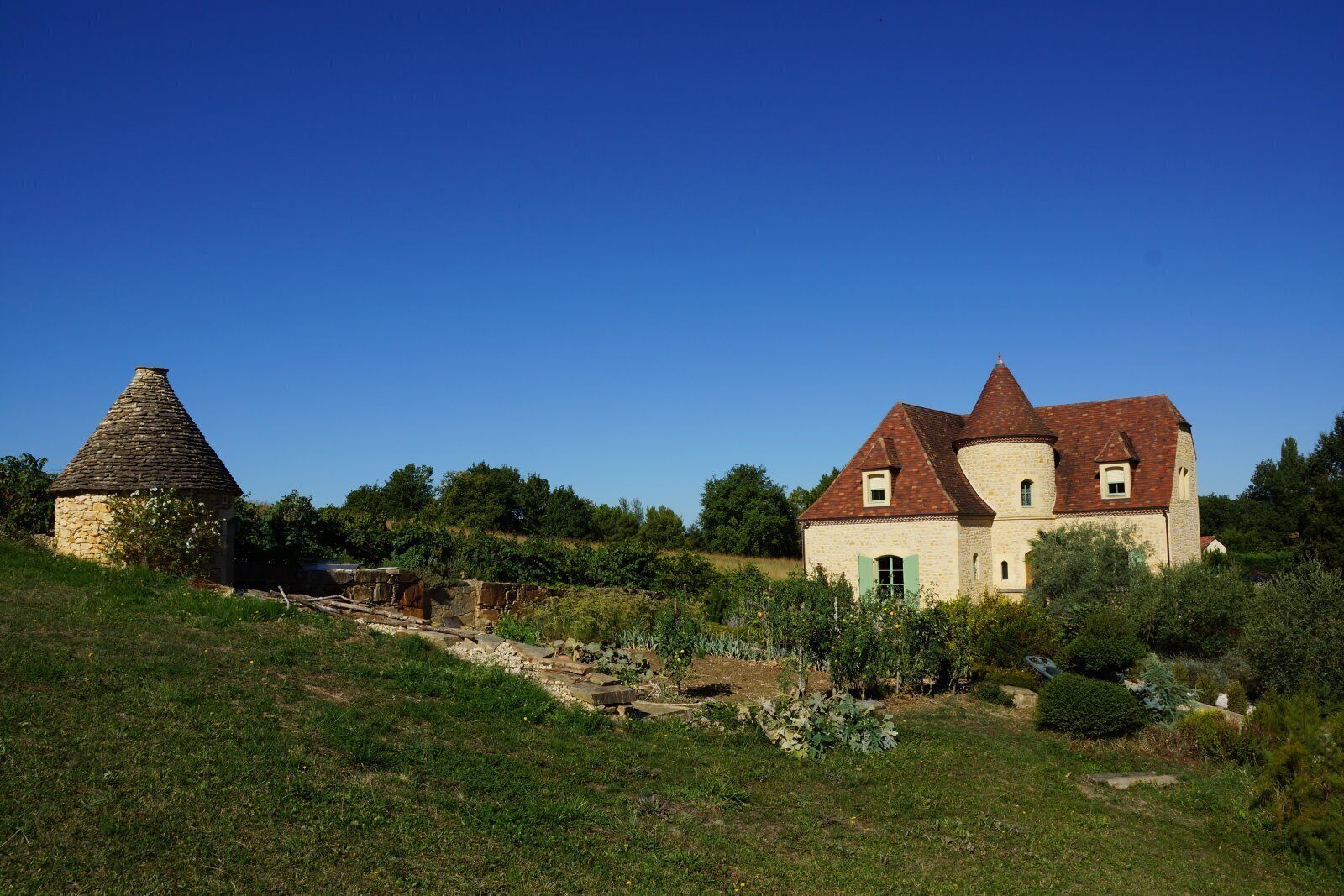 A stone house with a conical roof and a larger building with a pitched roof, surrounded by greenery and trees.