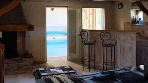 A rustic interior with exposed wooden beams, a stone fireplace, a large table with a patterned tablecloth, and a view of the sea through an open door. - Apartment with swimming pool and place of relaxation and leisure (Sarlat-la-Canéda)