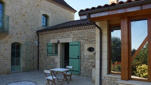 A stone building with a small patio area featuring a table and chairs, a wooden pergola, and a view of greenery. - Apartment with swimming pool and place of relaxation and leisure (Sarlat-la-Canéda)