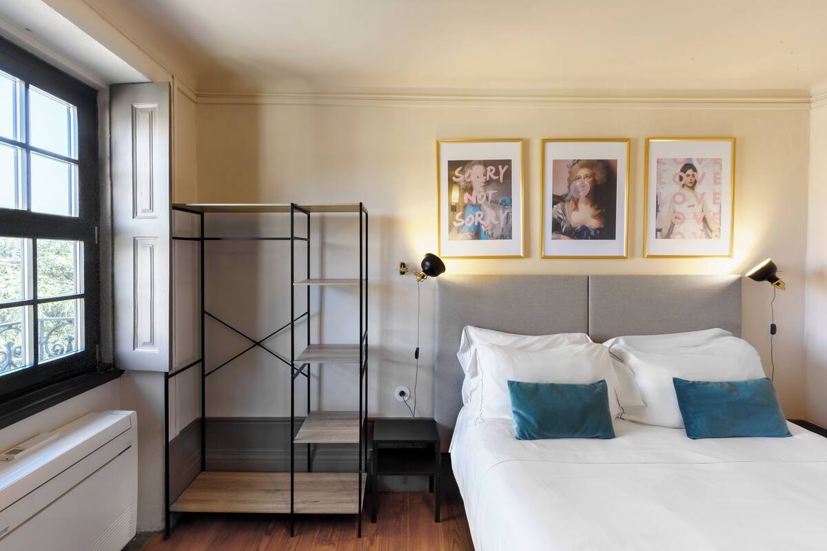 A bedroom with a bed, two pillows, a nightstand, a lamp, a shelf, and a window.