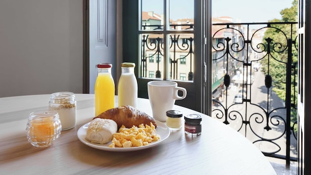 Uma mesa de café da manhã com croissants, ovos mexidos e uma xícara de café, com vista para uma varanda e prédios ao fundo.