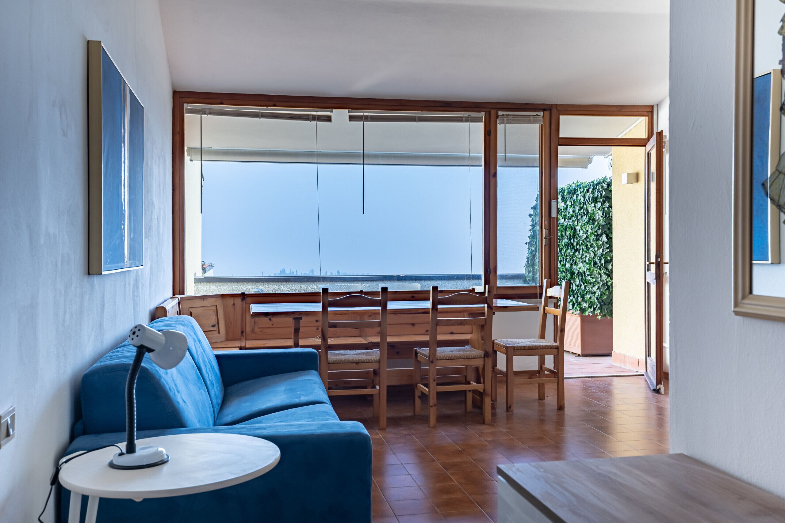 A room with a blue sofa, a small round table with a lamp, wooden chairs, and a large window with a view of the sea.