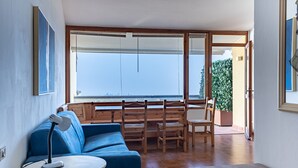 Een kamer met een blauwe bank, een klein rond tafeltje met een lamp, houten stoelen en een groot raam met uitzicht op de zee.