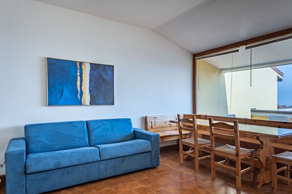 A room with a blue sofa, wooden dining table, and chairs, a large abstract painting on the wall, and a view through a large window.