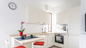 Cozinha moderna com armários brancos, bancada de madeira, pia e forno. Há um relógio na parede e um vaso de planta no balcão.