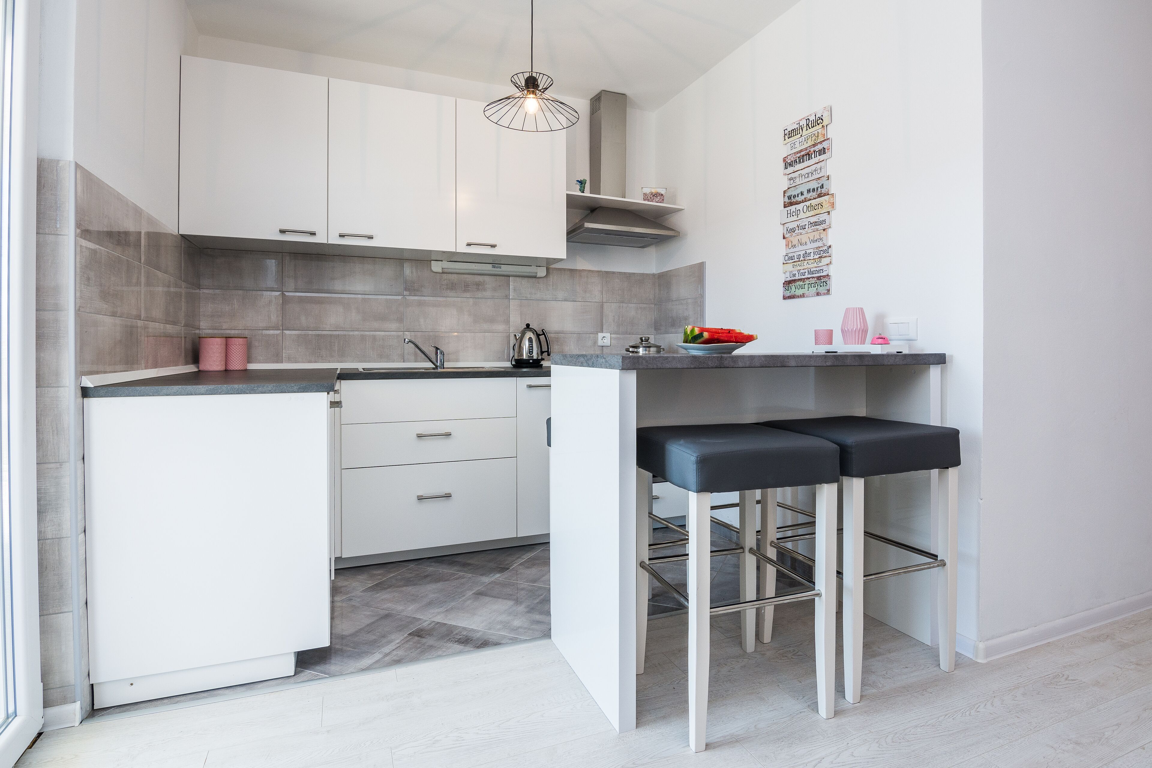 Een compacte keuken met witte kastjes, een grijze betegelde spatwand en een klein barretje met krukjes.