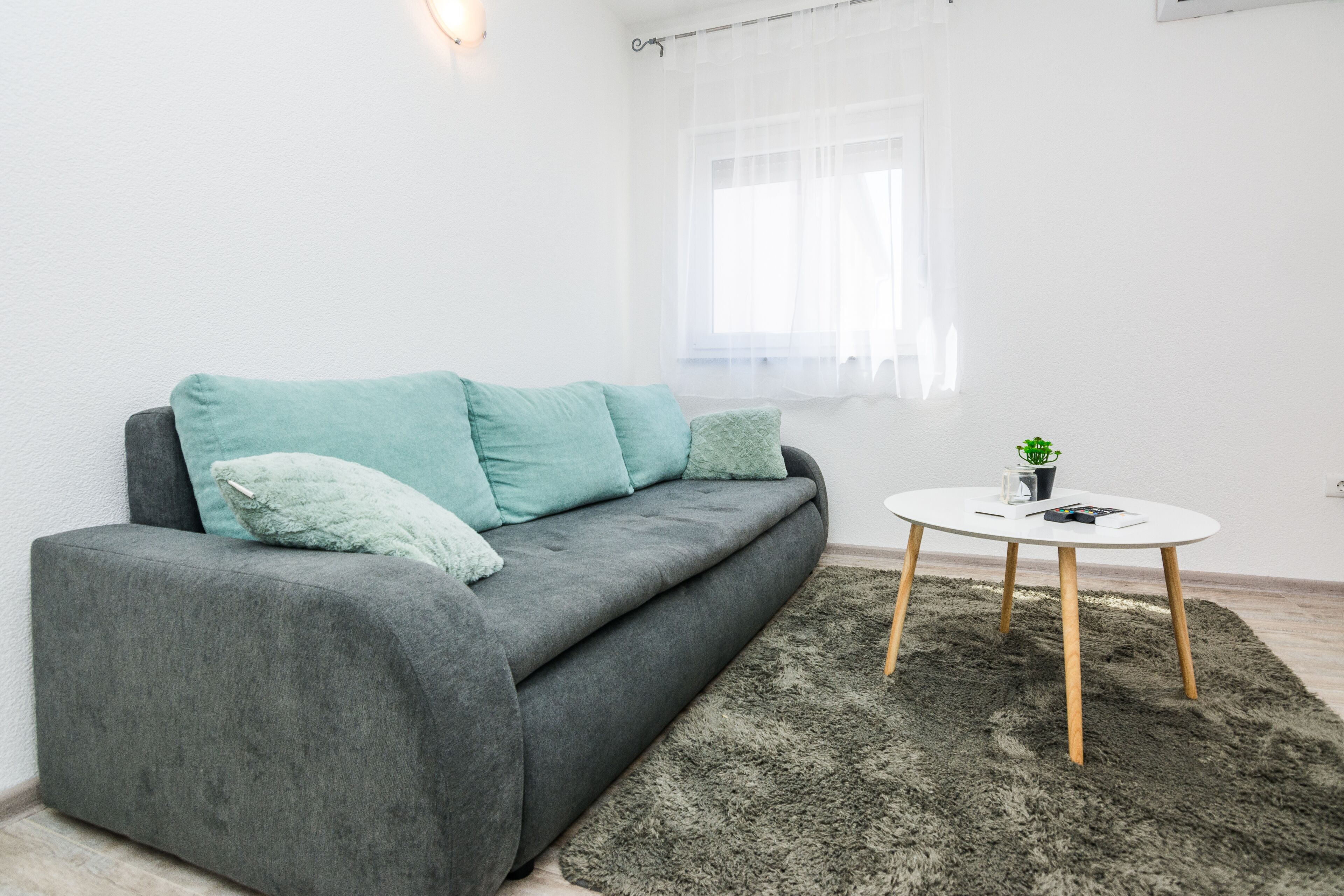 Uma sala de estar moderna com um sofá cinzento, almofadas verde-água, uma mesa de centro branca e redonda e uma pequena planta em vaso.