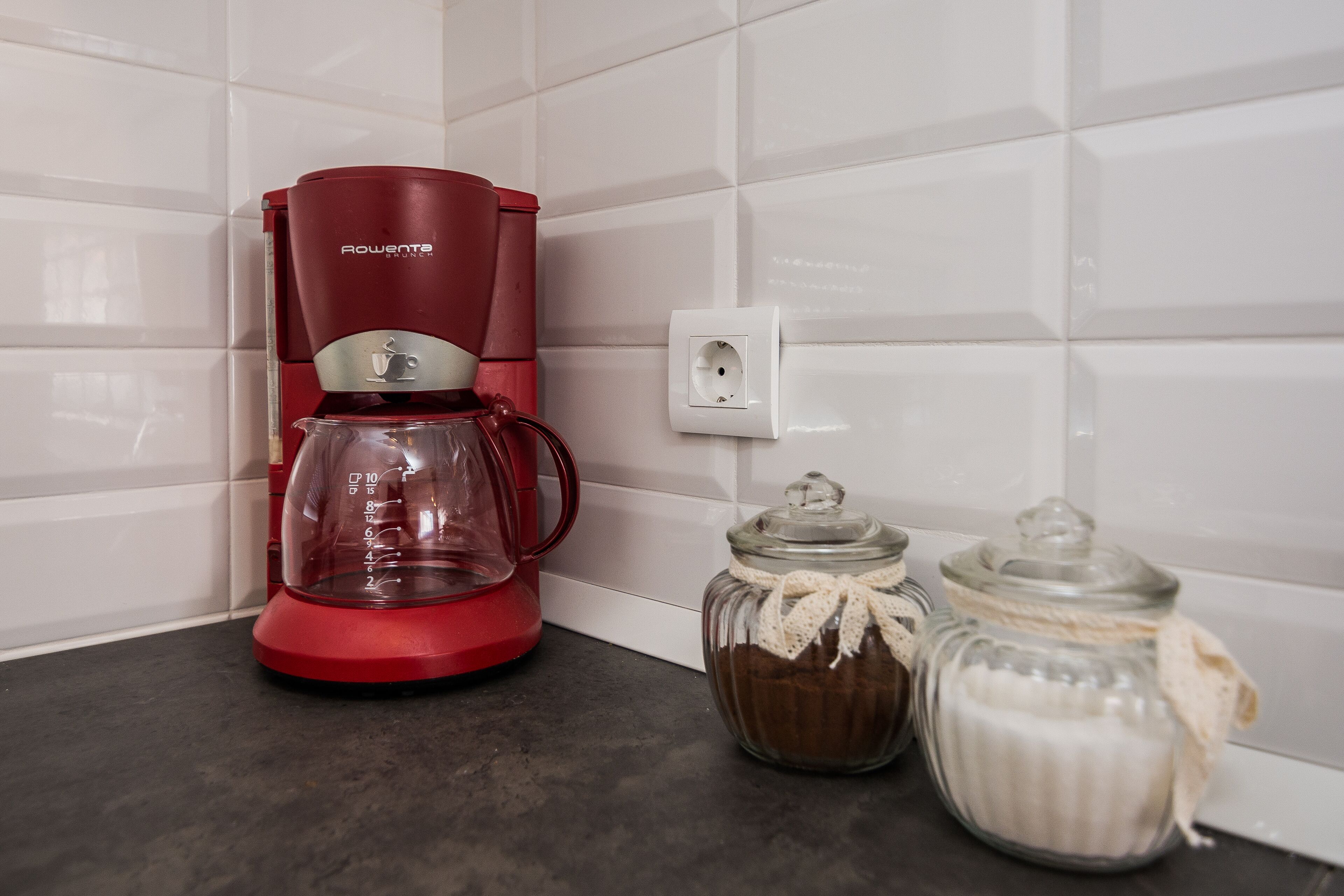 Een rode koffiezetapparaat, twee glazen potten met deksels, en een wit stopcontact op een aanrecht.
