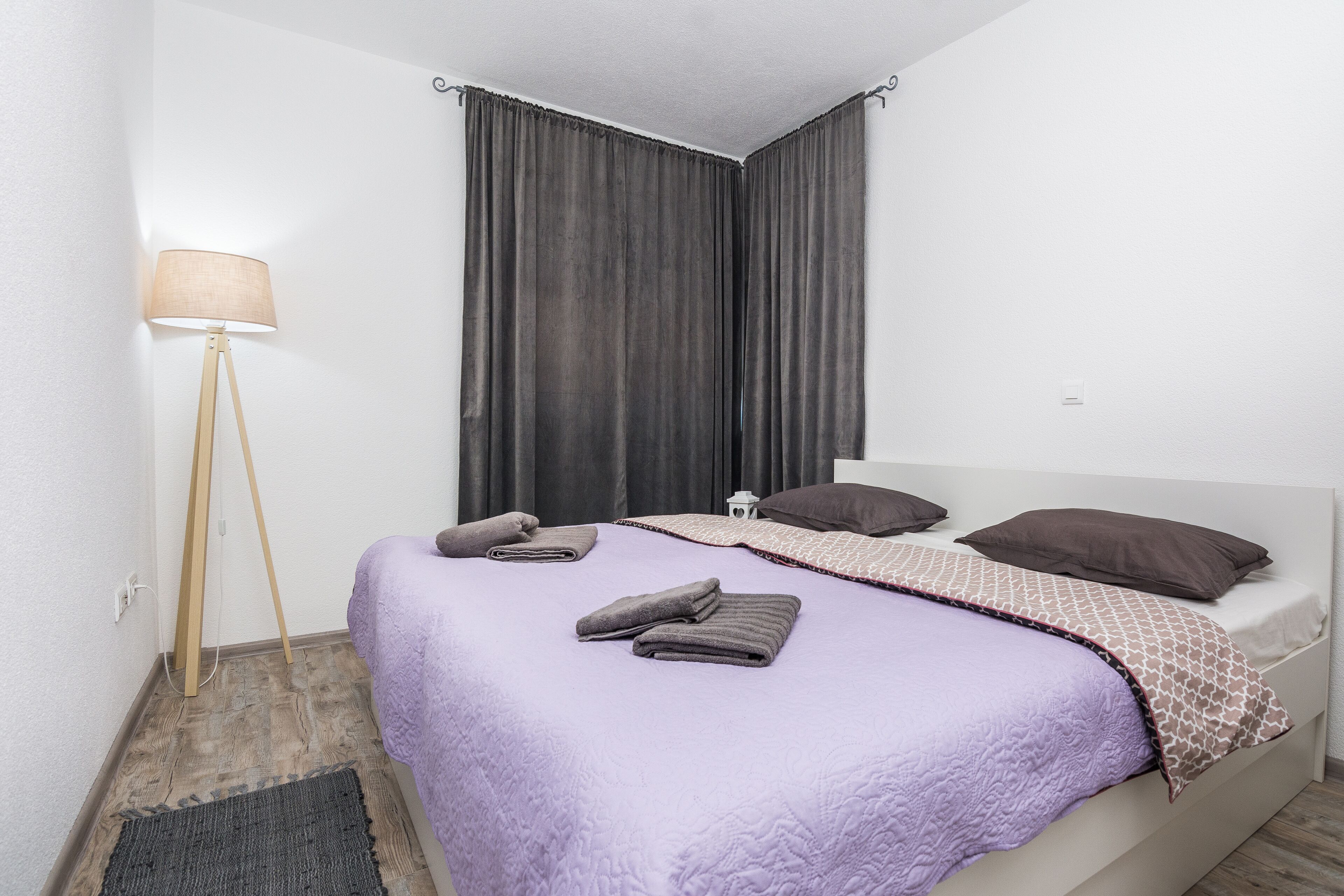 Un lit bien fait, recouvert d’une couverture violette, une lampe de chevet et une petite table avec une lampe.