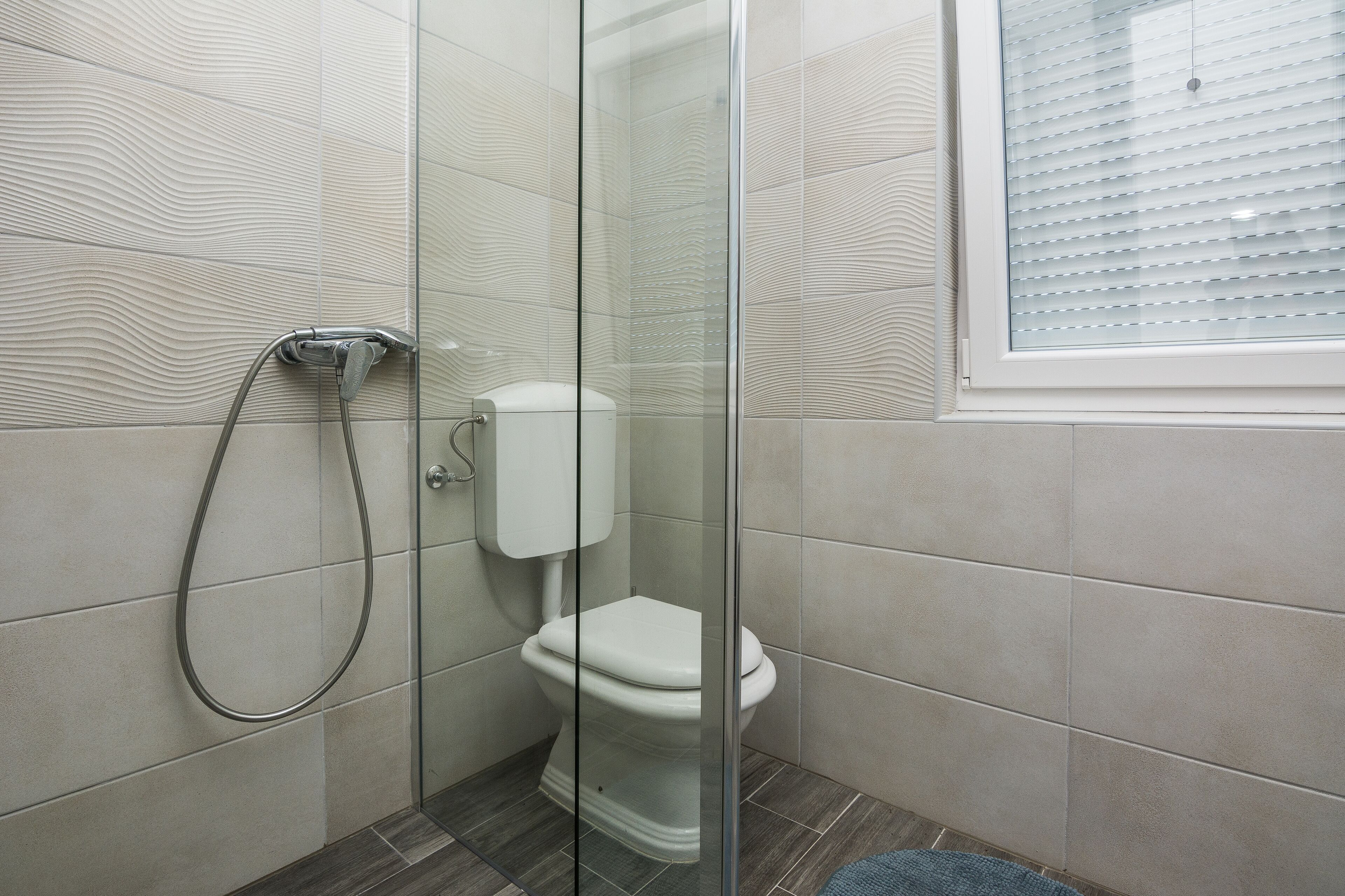 A modern bathroom with a glass shower enclosure, a toilet, and a window with blinds.