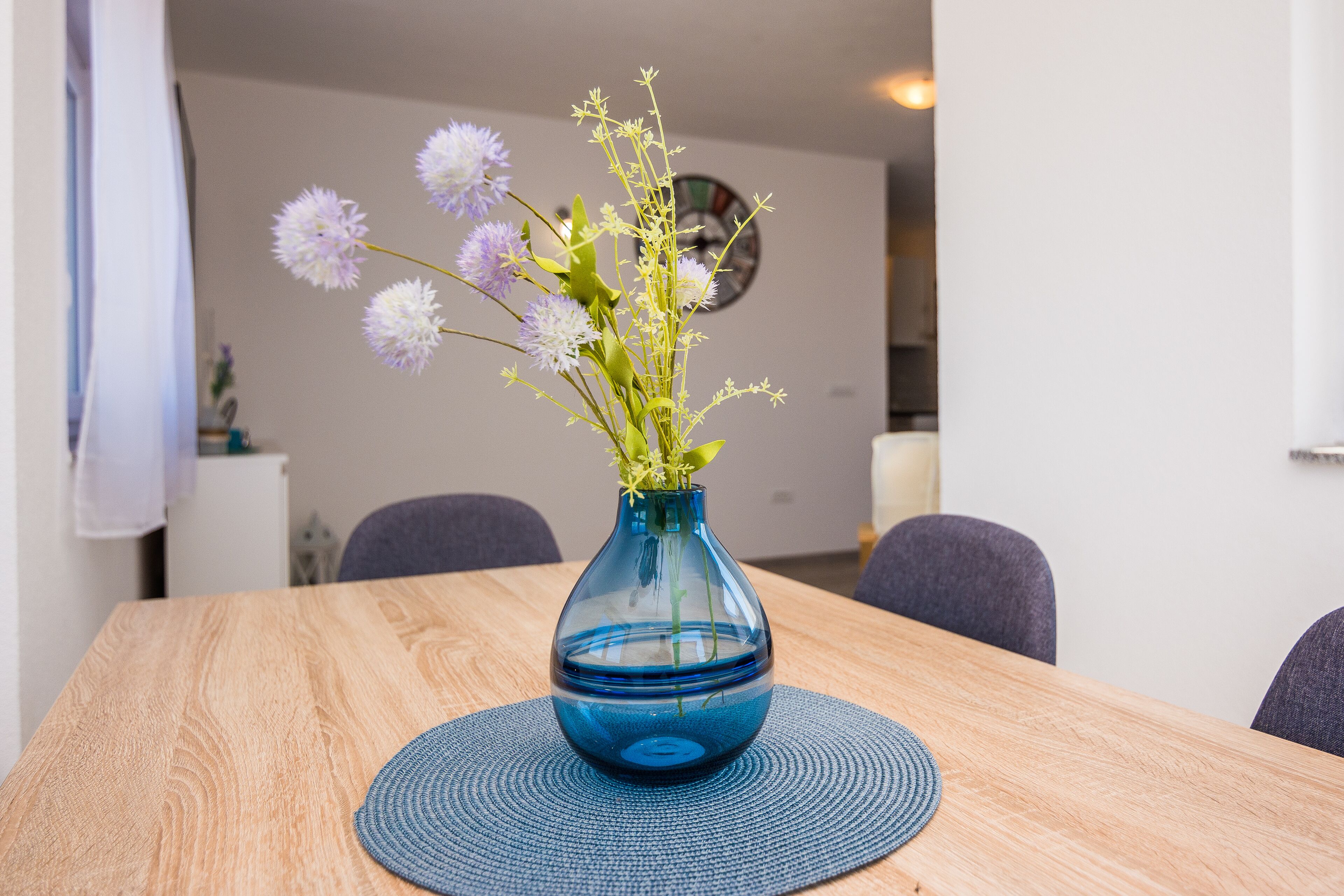 A dining table with a blue vase containing flowers, surrounded by chairs.
