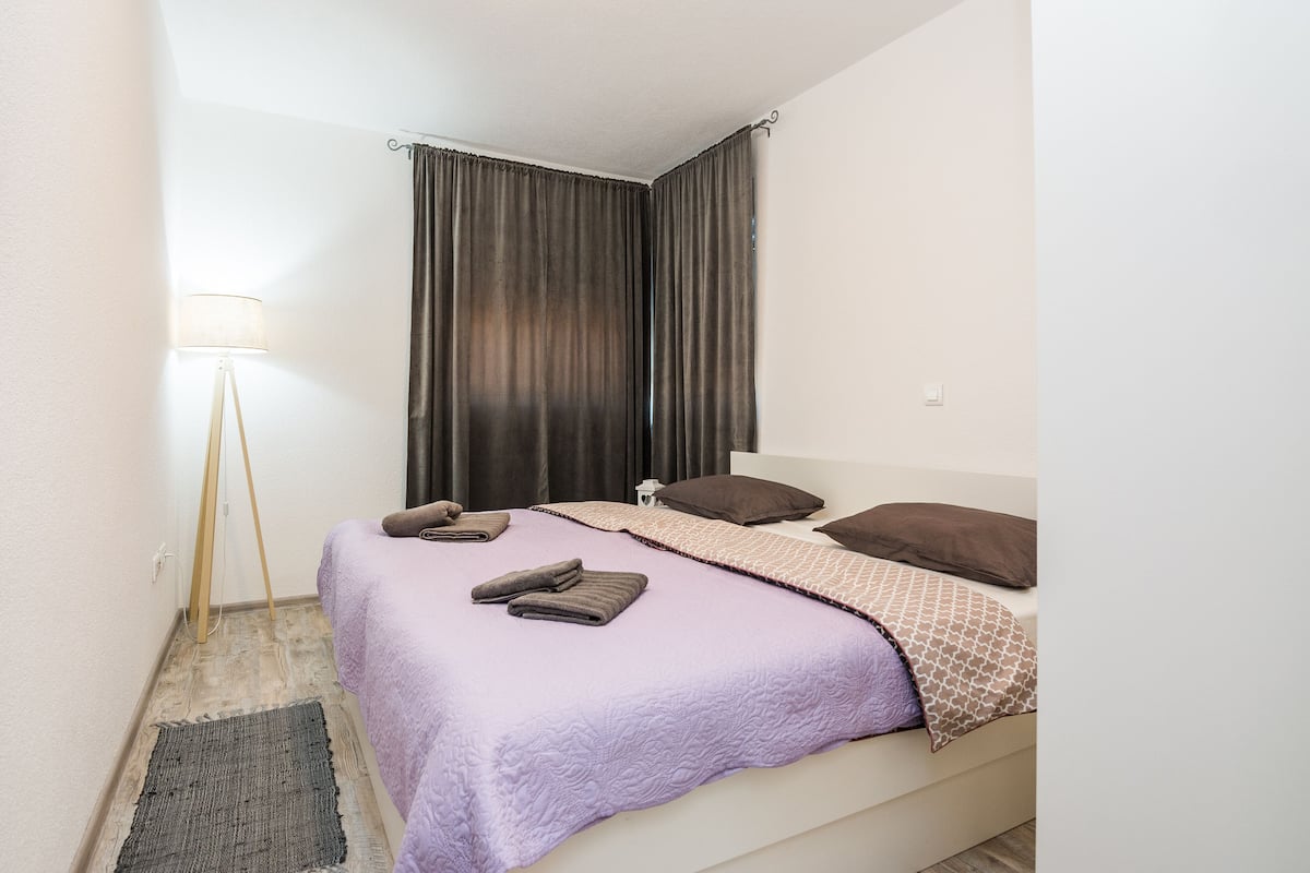 A neatly made bed with a purple blanket and pillows, a bedside lamp, and a dark curtain.