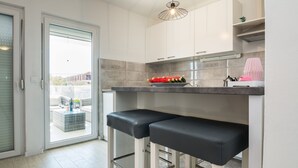 A modern kitchen with two bar stools, a countertop with a fruit bowl, and a view through a glass door. - Apartments Krpan (Vir)