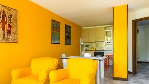 Ein hellgelbes Zimmer mit zwei gelben Stühlen, einem Glastisch und einer Küche im Hintergrund.