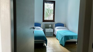 Una stanza piccola e luminosa con due letti singoli, una ventola a soffitto, una finestra vista verde e un termosifone.