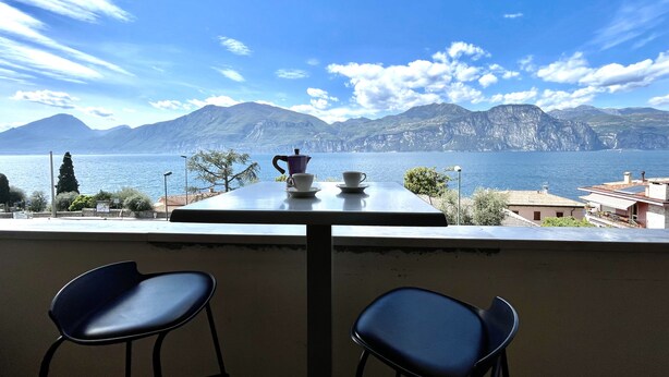 Un tavolo con una caffettiera e una tazza, due sedie e vista su un lago con montagne sullo sfondo.