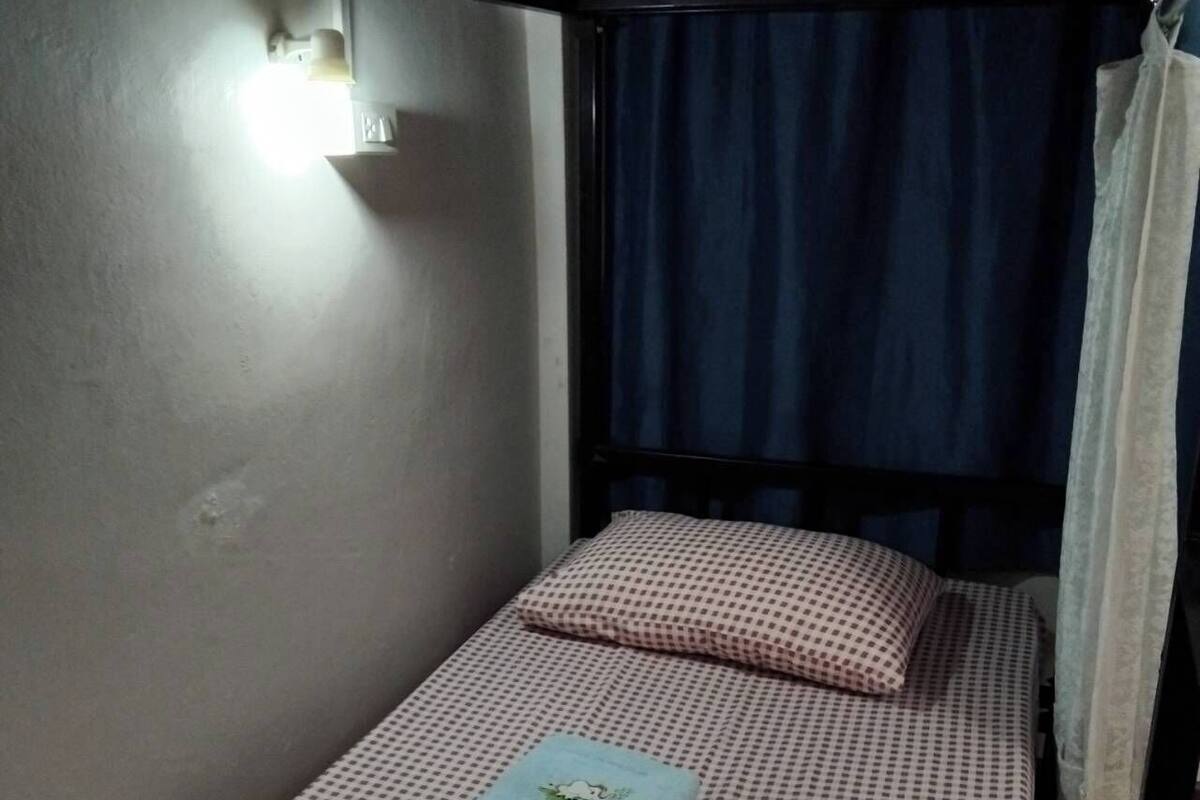 A small, single-bed room with a checkered bedspread, a folded towel, and a wall-mounted light.